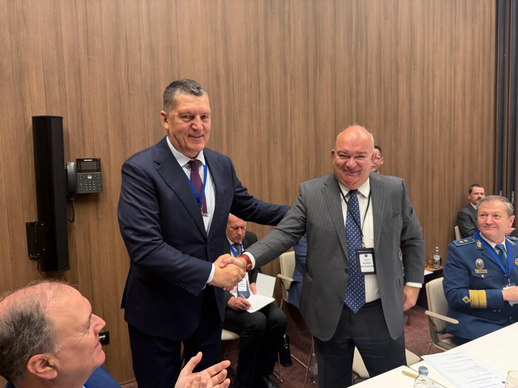 Zwei Teilnehmende in Anzugkleidung geben sich in einem Konferenzraum die Hand; weitere Teilnehmende sitzen im Hintergrund an einem Tisch mit Unterlagen und Namensschildern.