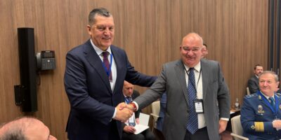 Zwei Teilnehmende in Anzugkleidung geben sich in einem Konferenzraum die Hand; weitere Teilnehmende sitzen im Hintergrund an einem Tisch mit Unterlagen und Namensschildern.