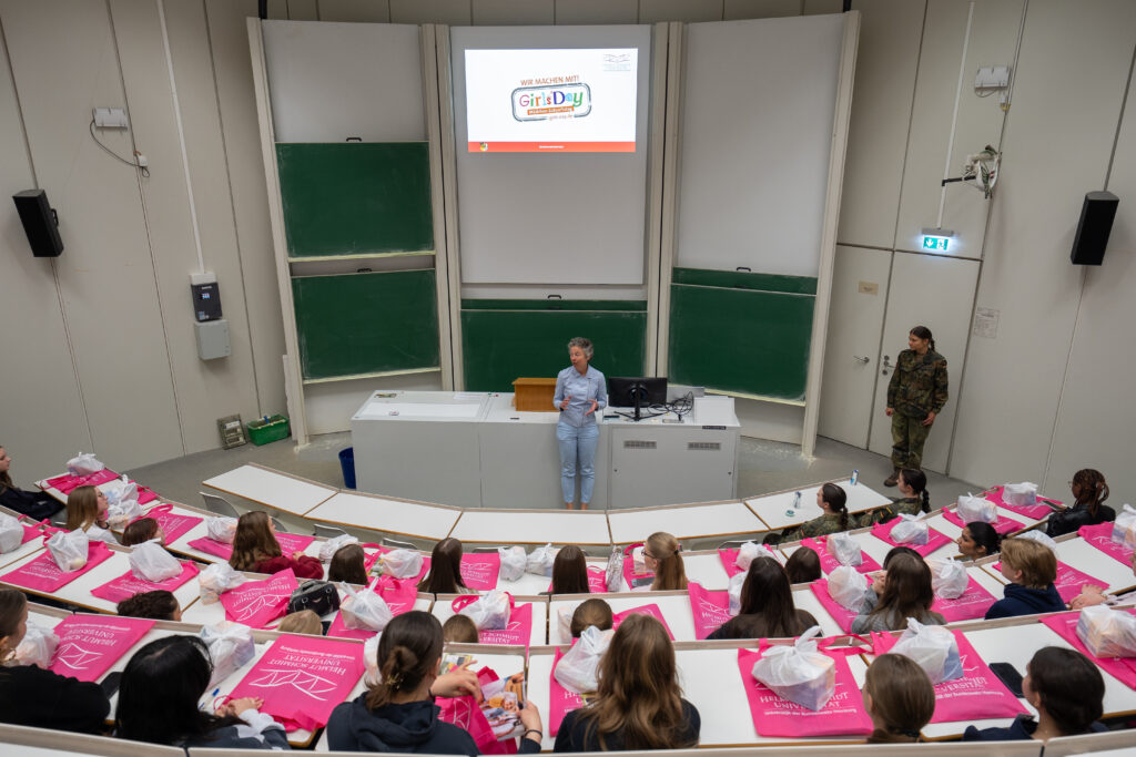 Blick von hinten in einen Hörsaal: Schülerinnen sitzen an Reihenplätzen, vorne spricht eine Person, auf der Leinwand ist eine Präsentation zum Girls’ Day zu sehen.