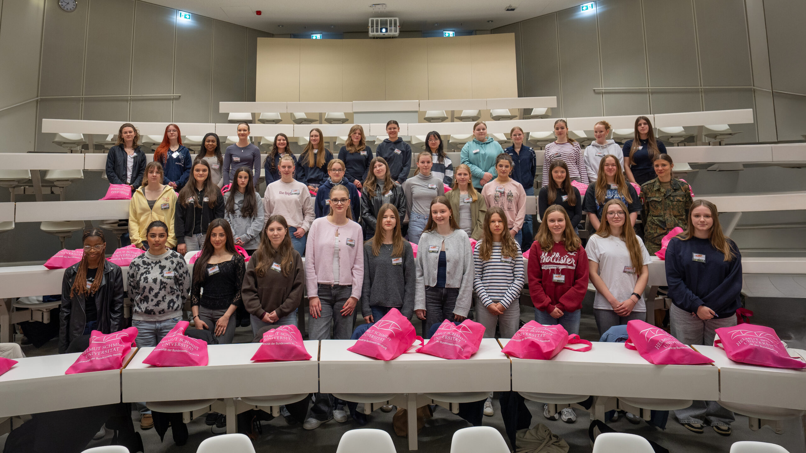 Große Gruppe von Schülerinnen steht in einem Hörsaal der Helmut‑Schmidt‑Universität/Universität der Bundeswehr Hamburg, vor ihnen liegen pinke Stofftaschen mit Universitätslogo.