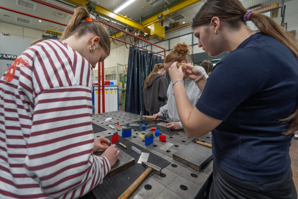 Schülerinnen arbeiten an einer Werkbank und schlagen mit Metallbuchstaben Namen in kleine Metallschilder ein.
