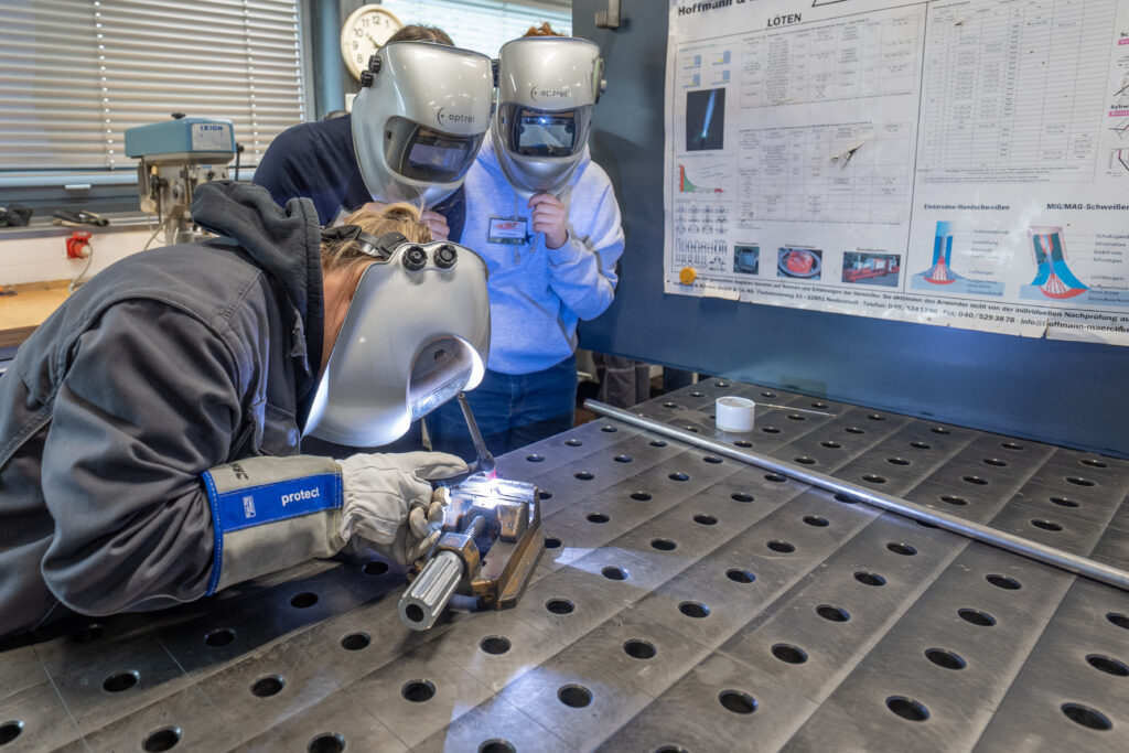 Schülerinnen führen Lötarbeiten an einem Metallbauteil in einer Werkstatt aus, sie tragen Schutzkleidung und arbeiten an einer stabilen Werkbank.