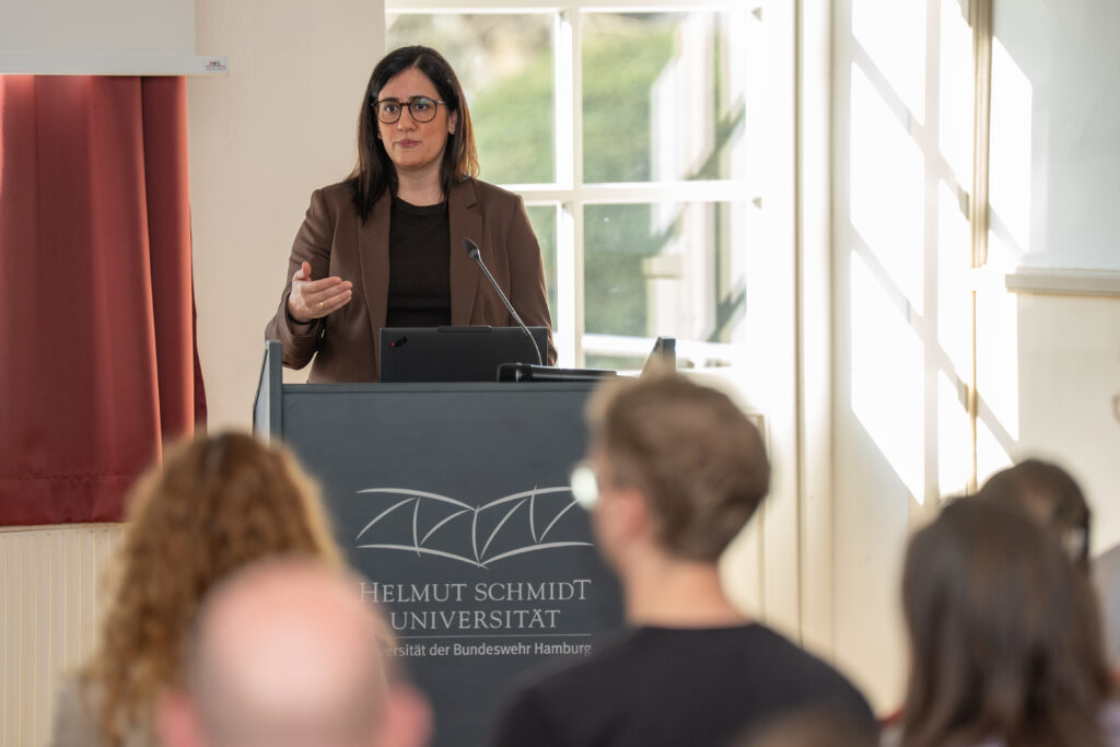 Eine Frau steht an einem Rednerpult mit HSU/UniBw H Logo und spricht vor einem Publikum. 