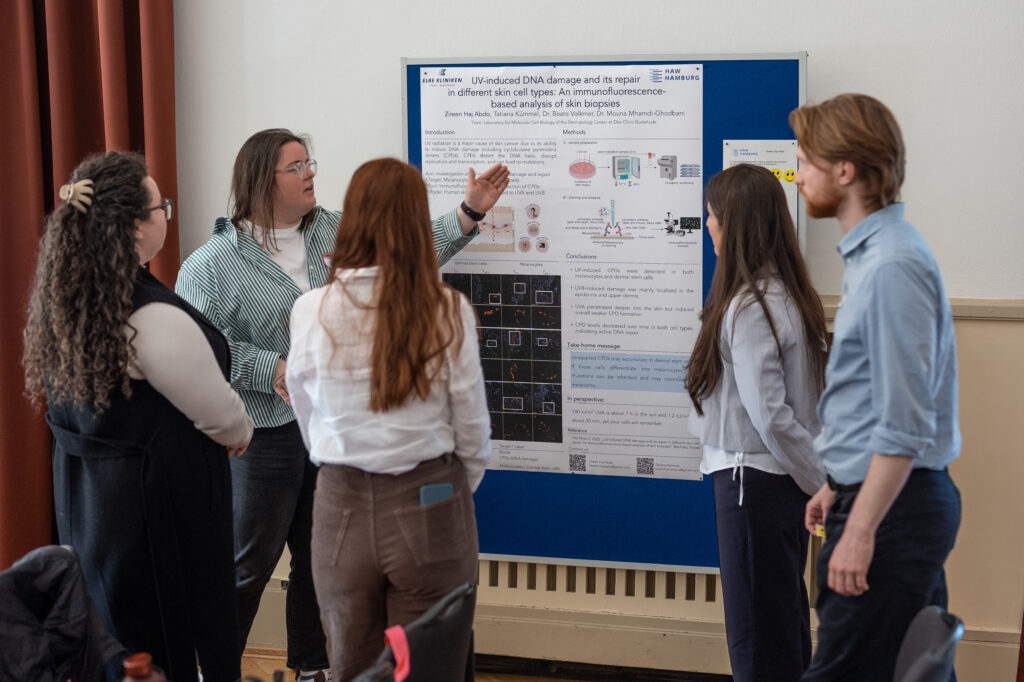 Vier Personen stehen vor einem wissenschaftlichen Poster. Dieses wird ihnen angeregt von einer Person vorgestellt. 