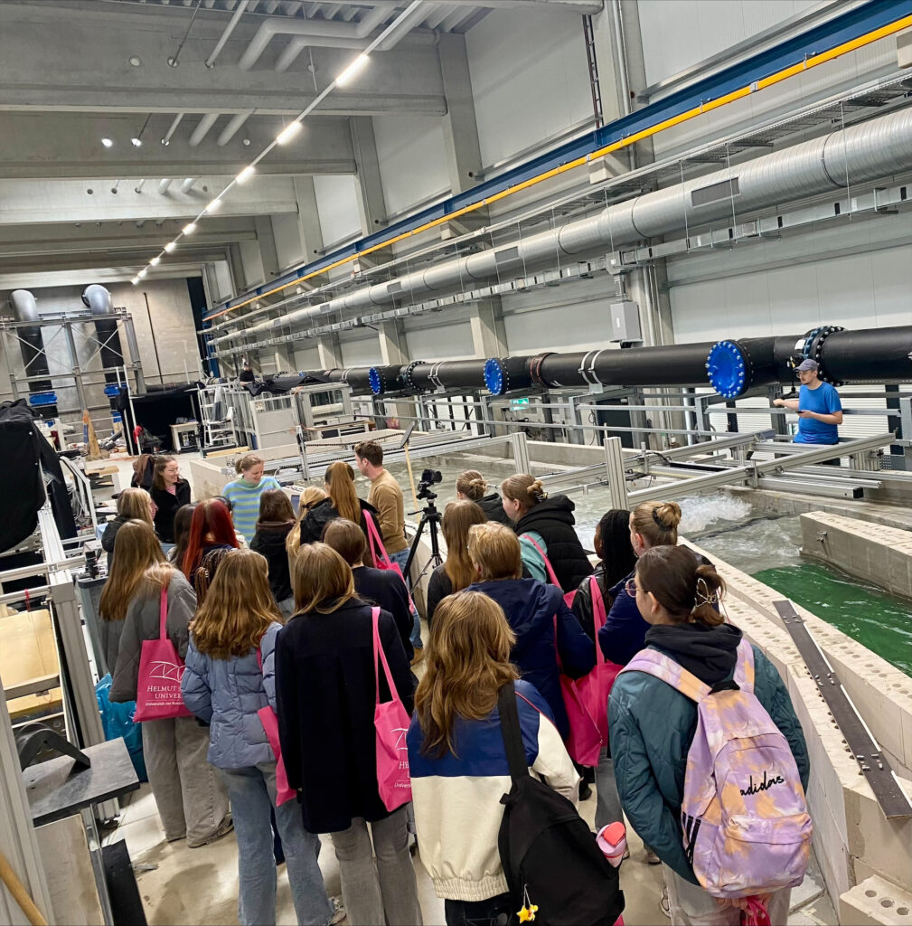 Gruppe von Schülerinnen steht in einem großen Wasserbaulabor vor einem Strömungsmodell mit Wasserkanal und technischen Anlagen.