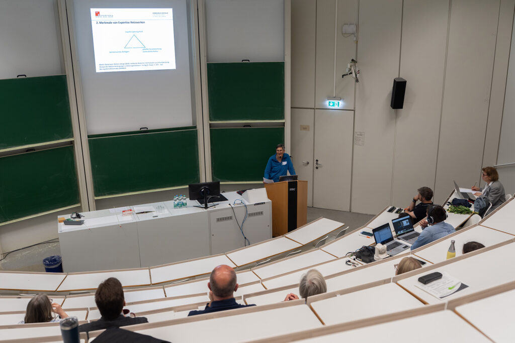 Keynote‑Vortrag im Hörsaal: Eine Referentin spricht am Rednerpult vor einer Präsentationsfolie zu Expertise Netzwerken in der Hochschullehre.