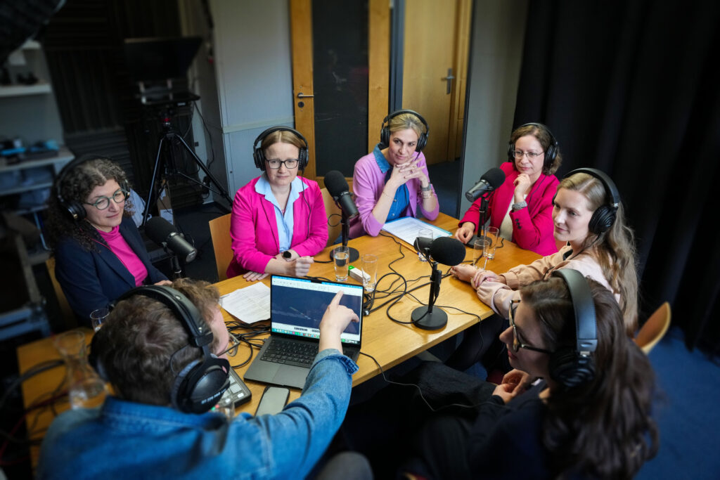 Die Podcast-Hostinnen sitzen mit Kopfhörern um einen Tisch in einem Podcast‑Studio und diskutieren mittels Mikrofonen gemeinsam.