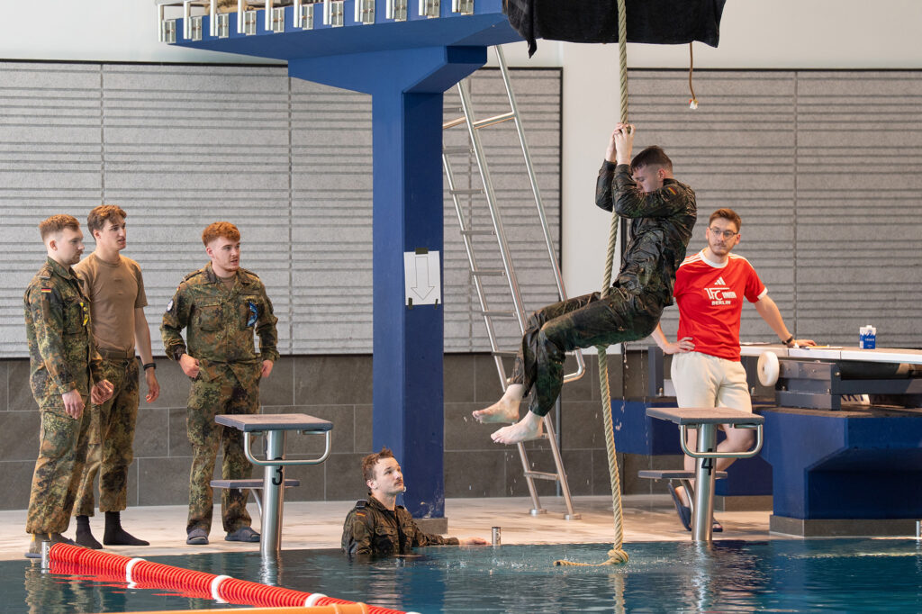 Mehrere Personen in Sport- und Dienstkleidung beobachten, wie eine Person an einem Seil über einem Schwimmbecken nach oben klettert.