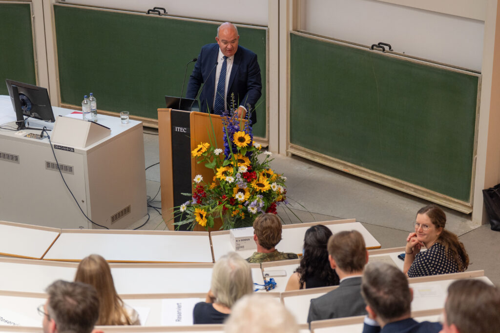 Eine Person steht an einem Rednerpult in einem Hörsaal der HSU/UniBw H und spricht vor Publikum; vor dem Pult befindet sich ein Blumenarrangement.