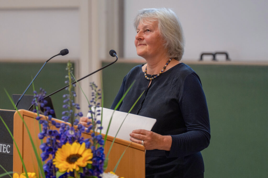 Eine Person steht an einem Rednerpult mit Mikrofonen in einem Hörsaal und hält Unterlagen in der Hand; im Vordergrund ist Blumenschmuck zu sehen.