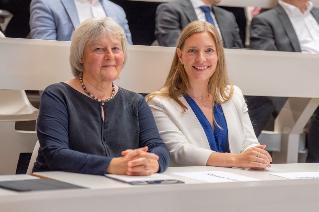 Zwei Personen sitzen nebeneinander in einem Hörsaal der HSU/UniBw H und blicken in die Kamera; im Hintergrund sind weitere Teilnehmende zu sehen.