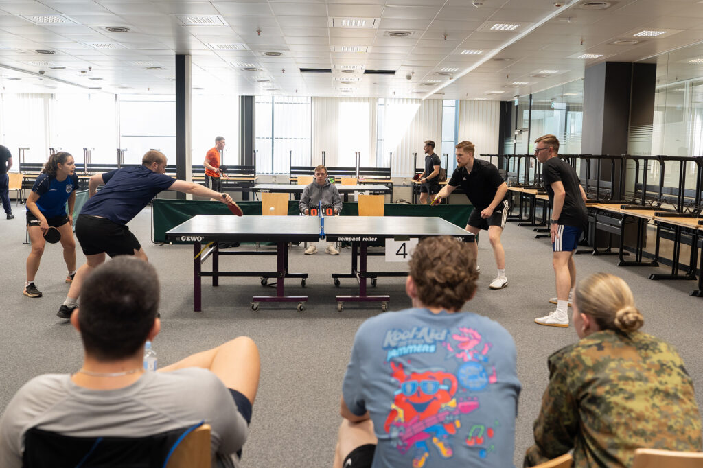 Mehrere Personen spielen Tischtennis an einer Platte, während andere sitzend im Vordergrund zuschauen.
