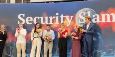 Dr. Babis C. Karpouchtsis steht lachend und mit enem Blumenstrauß auf der Bühne des Security Slams 2026. Um ihn herum stehen weitere Teilnehmende.