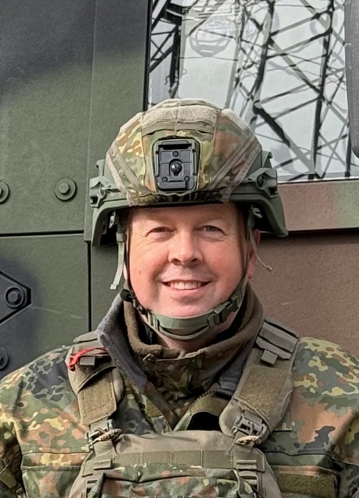 Person in Flecktarnuniform mit Schutzausrüstung steht vor technischem Hintergrund.