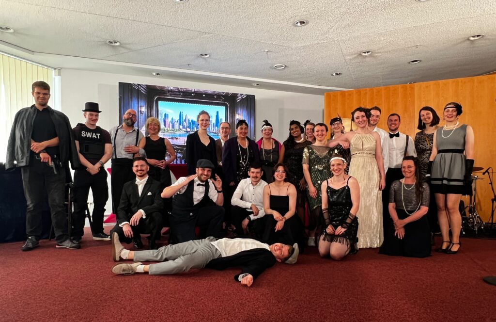 Gruppe von rund 25 Mitwirkenden eines Musicals in 1920er‑Jahre‑Kostümen steht und sitzt gemeinsam auf einer Bühne; im Vordergrund liegt eine Person als Teil der Szene.