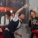 Mehrere Darstellende an einem Stehtisch agieren miteinander; eine Person hebt eine Flasche, andere halten Gläser und Requisiten.