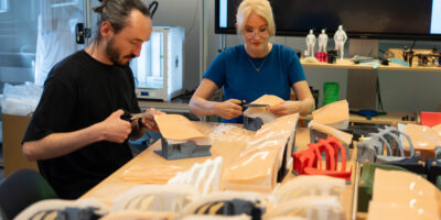 Zwei Personen schneiden im Labor Silikonmaterial für Trainingsmodelle; auf dem Tisch liegen mehrere Modellbauteile.