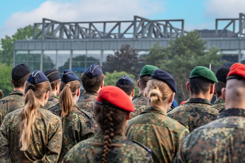 Mehrere Personen in Flecktarnuniform stehen auf einem Exerzier- oder Sportplatz und blicken von hinten in dieselbe Richtung. Einige tragen verschiedenfarbige Baretts, darunter rote, grüne und dunkelblaue. Im Hintergrund ist ein modernes Gebäude mit Glasfassade und Stahlkonstruktion zu sehen.