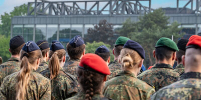 Mehrere Personen in Flecktarnuniform stehen auf einem Exerzier- oder Sportplatz und blicken von hinten in dieselbe Richtung. Einige tragen verschiedenfarbige Baretts, darunter rote, grüne und dunkelblaue. Im Hintergrund ist ein modernes Gebäude mit Glasfassade und Stahlkonstruktion zu sehen.
