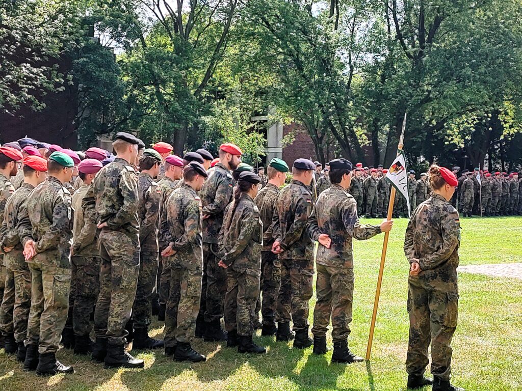 Gruppe von Studierenden in Feldanzügen steht auf einer Grünfläche in Formation. Im Vordergrund hält eine Person eine Fahne, weitere Teilnehmende sind im Hintergrund zwischen Bäumen versammelt.
