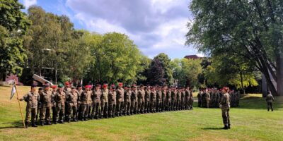 Gruppe von Studierenden in Feldanzügen steht auf einer Grünfläche in Formation. Im Vordergrund hält eine Person eine Fahne, weitere Teilnehmende sind im Hintergrund zwischen Bäumen versammelt.