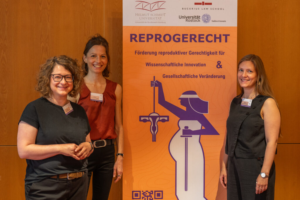 Drei Personen stehen vor einem orangefarbenen Banner des Netzwerks ReproGerecht mit den Logos der HSU/UniBw H, der Bucerius Law School und der Universität Rostock. In der Mitte steht Prof. Dr. Henrike von Scheliha, rechts Prof. Dr. Dana-Sophia Valentiner.