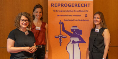 Drei Personen stehen vor einem orangefarbenen Banner des Netzwerks ReproGerecht mit den Logos der HSU/UniBw H, der Bucerius Law School und der Universität Rostock. In der Mitte steht Prof. Dr. Henrike von Scheliha, rechts Prof. Dr. Dana-Sophia Valentiner.