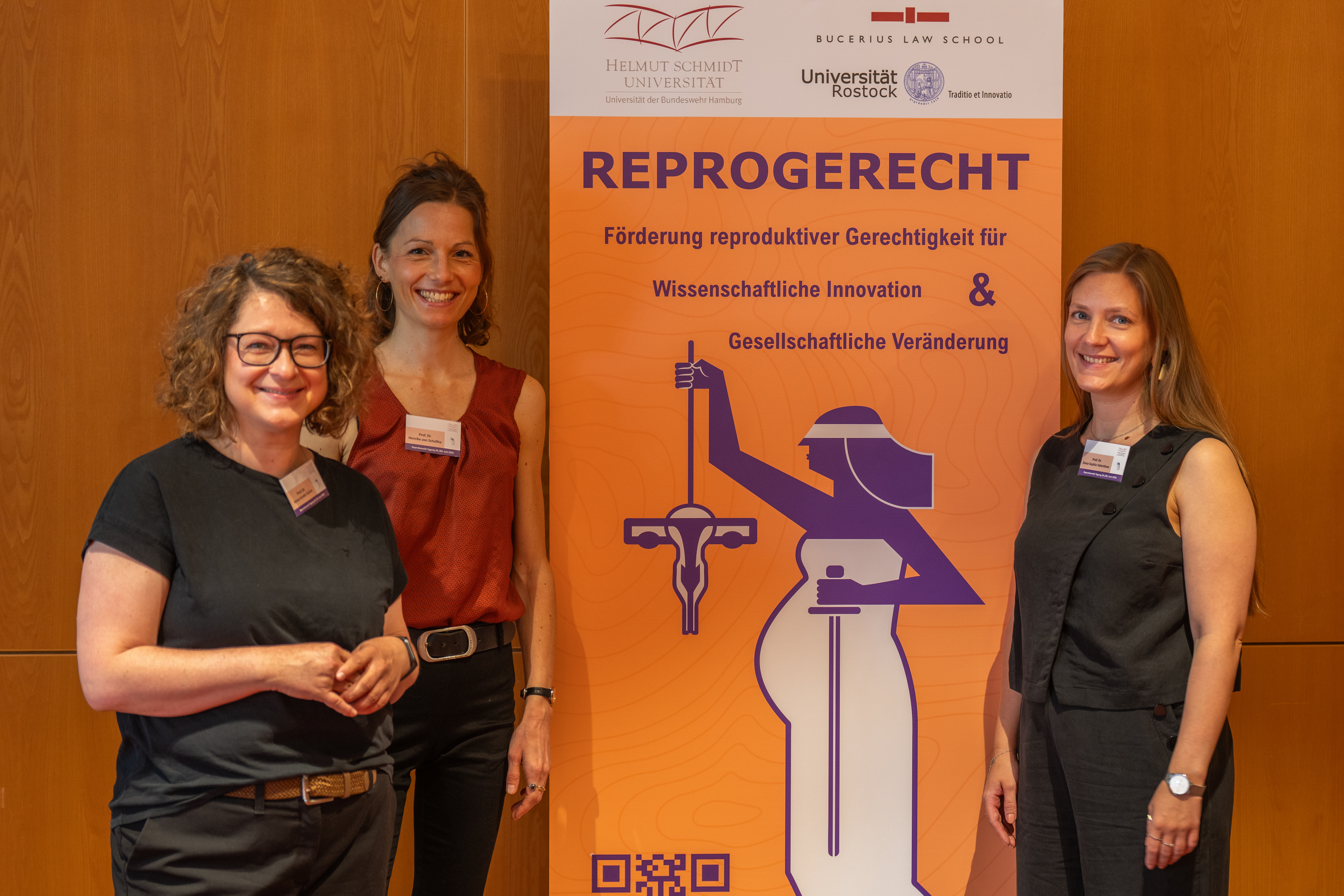 Drei Personen stehen vor einem orangefarbenen Banner des Netzwerks ReproGerecht mit den Logos der HSU/UniBw H, der Bucerius Law School und der Universität Rostock. In der Mitte steht Prof. Dr. Henrike von Scheliha, rechts Prof. Dr. Dana-Sophia Valentiner.