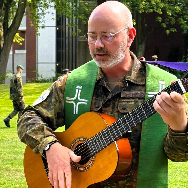 Person in Feldanzug mit grünem liturgischem Stola spielt im Freien Gitarre während eines Feldgottesdienstes. Im Hintergrund sind weitere Angehörige der Bundeswehr auf einer Rasenfläche zu sehen.