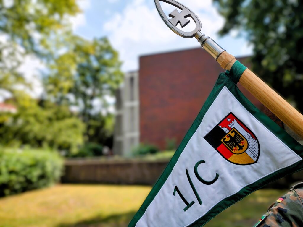 Nahaufnahme eines Wimpels mit Bundesadler und der Aufschrift „1/C“ an einer Holzstange. Im Hintergrund sind Bäume und ein Gebäude auf dem Campus unscharf zu erkennen.