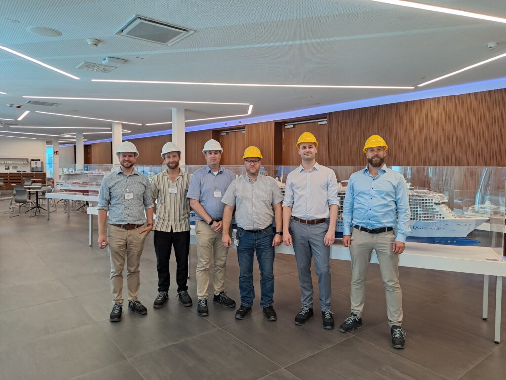 Sechs Teilnehmende des Forschungsprojekts H2MASS stehen während eines Kick-off-Workshops in einer Ausstellungshalle der MEYER WERFT vor mehreren Modellen von Passagierschiffen. Einige der Beteiligten tragen Schutzhelme. Im Hintergrund sind Vitrinen mit Schiffsmodellen zu sehen.