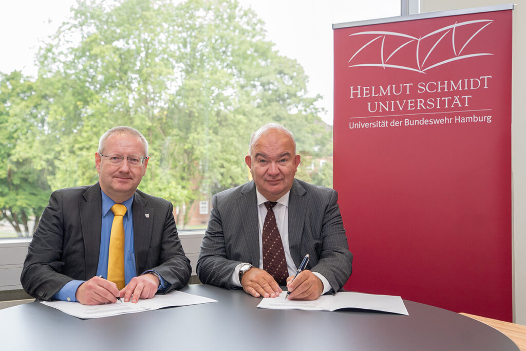 Zie Männer sitzen im Anzug vor einem Hellmut-Schmidt-Universität Poster. Vor ihnen liegt Papier und sie halten Stifte in der Hand.