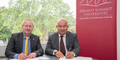 Zie Männer sitzen im Anzug vor einem Hellmut-Schmidt-Universität Poster. Vor ihnen liegt Papier und sie halten Stifte in der Hand.