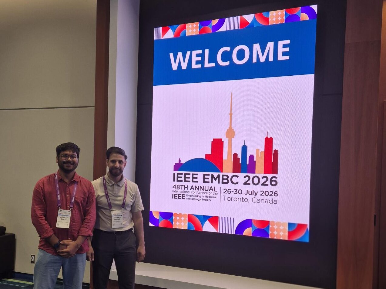 Zwei wissenschaftliche Mitarbeiter der Professur für Data Engineering stehen vor einer digitalen Anzeigetafel der IEEE Engineering in Medicine and Biology Conference (EMBC) 2026 in Toronto. Die Konferenzanzeige zeigt das Logo der Veranstaltung sowie Veranstaltungsort und -datum.