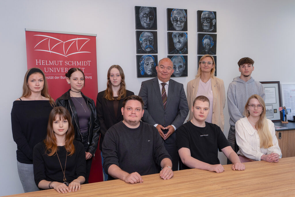 Gruppenfoto der neuen Auszubildenden des Jahrgangs 2026 gemeinsam mit dem Präsidenten der HSU/UniBw H in einem Besprechungsraum der Universität. Die Gruppe steht und sitzt vor einem Roll-up der Universität.