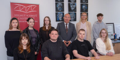 Gruppenfoto der neuen Auszubildenden des Jahrgangs 2026 gemeinsam mit dem Präsidenten der HSU/UniBw H in einem Besprechungsraum der Universität. Die Gruppe steht und sitzt vor einem Roll-up der Universität.