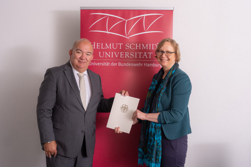 Zwei Personen stehen vor einem Roll-up der Helmut-Schmidt-Universität/Universität der Bundeswehr Hamburg und halten gemeinsam eine Ernennungsurkunde.