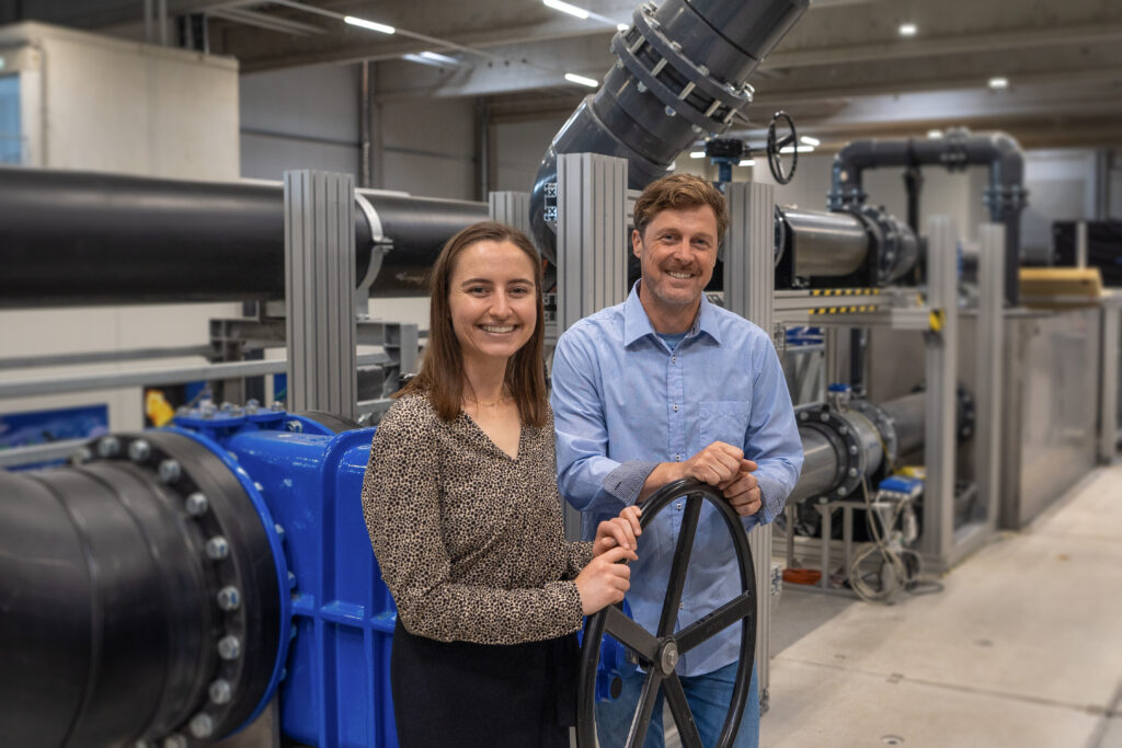 Zwei Personen stehen vor einer großmaßstäblichen wasserbaulichen Versuchsanlage in der Wasserbauhalle der HSU/UniBw Hamburg. Im Hintergrund sind Rohrleitungen, Armaturen und technische Komponenten der Forschungsinfrastruktur zu sehen.