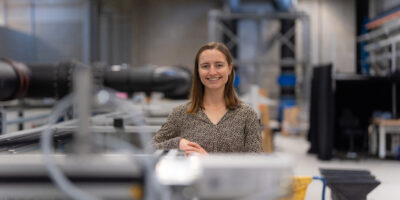 Portrait von Thea Giliomee in der Wasserbauhalle der HSU/UniBw Hamburg. Im Hintergrund sind Versuchsanlagen und Rohrleitungen für hydraulische Untersuchungen zu erkennen.