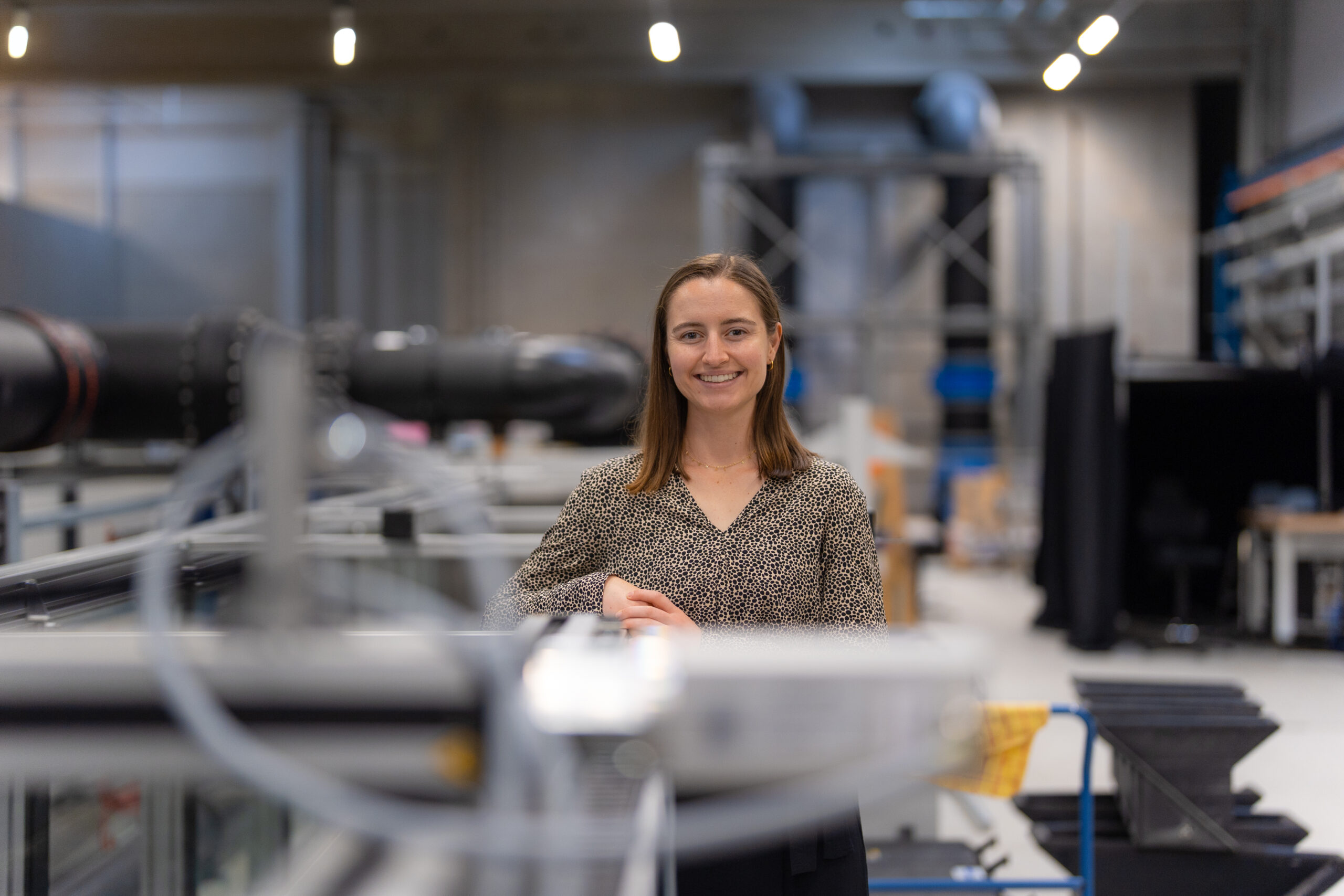Portrait von Thea Giliomee in der Wasserbauhalle der HSU/UniBw Hamburg. Im Hintergrund sind Versuchsanlagen und Rohrleitungen für hydraulische Untersuchungen zu erkennen.