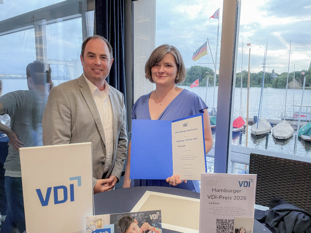 Prof. Dr.-Ing. Julian Jepsen und Oberleutnant Sarah stehen mit der Urkunde des VDI-Preises 2026 vor einer Fensterfront mit Blick auf die Außenalster. Auf einem Tisch im Vordergrund sind weitere Unterlagen und ein Hinweis auf den Hamburger VDI-Preis zu sehen.