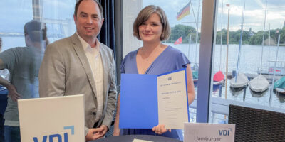 Prof. Dr.-Ing. Julian Jepsen und Oberleutnant Sarah stehen mit der Urkunde des VDI-Preises 2026 vor einer Fensterfront mit Blick auf die Außenalster. Auf einem Tisch im Vordergrund sind weitere Unterlagen und ein Hinweis auf den Hamburger VDI-Preis zu sehen.