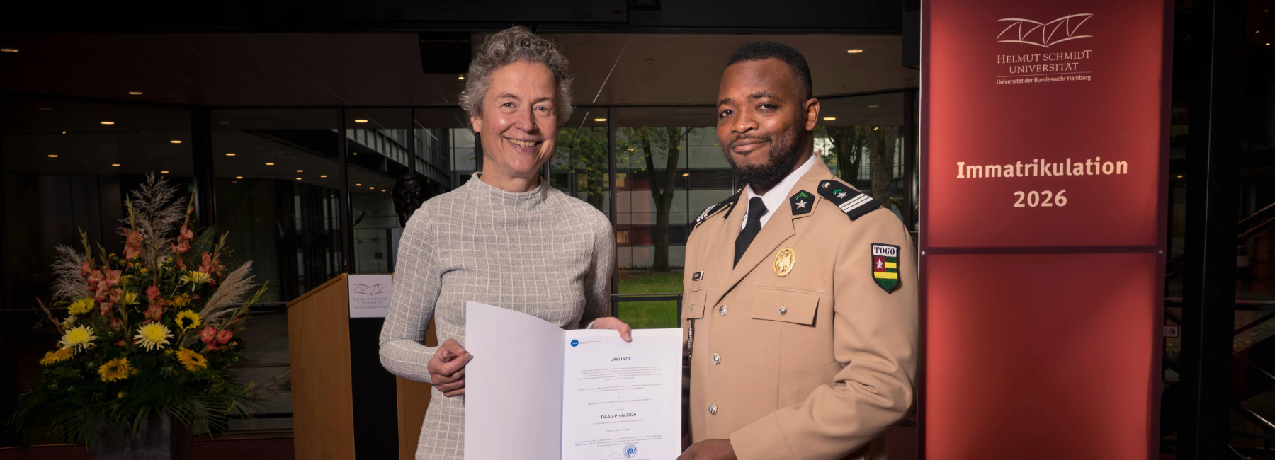Two people are standing side by side at the 2026 enrollment ceremony, holding the certificate for the 2026 DAAD Prize up to the camera. In the background, a red HSU/UniBw H stele bearing the inscription “Enrollment 2026” is visible.
