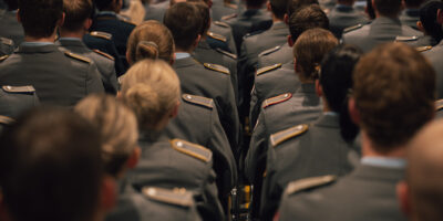 Mehrere Reihen von Studierenden in Uniform sitzen um den Roten Platz und verfolgen die Immatrikulationsfeier der HSU/UniBw H. Die Aufnahme zeigt das Publikum von hinten.