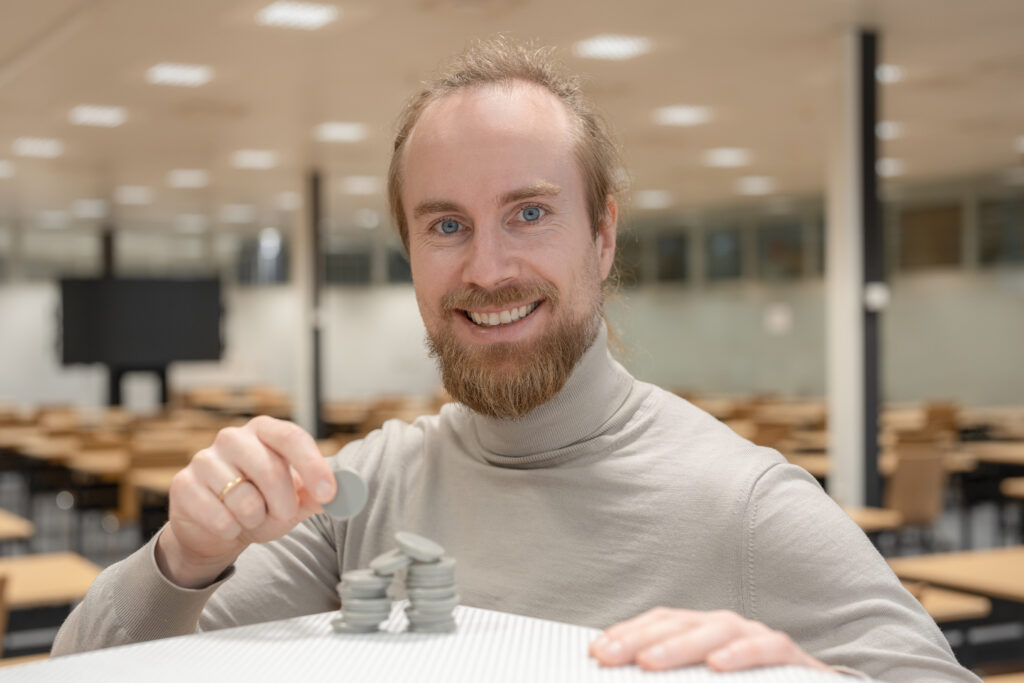 Eine Person sitzt an einem Tisch vor mehreren kleinen Stapeln aus runden, grauen Spielsteinen und hält einen weiteren Stein über die Stapel. Im Hintergrund sind Tische und Stühle eines hellen Innenraums zu sehen.